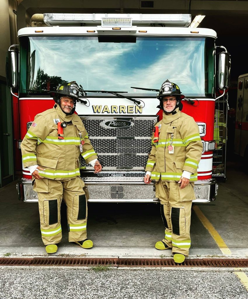 Two firefighters in full gear stand in front of a red fire truck labeled "Warren." Both are smiling and posing with one hand on the truck grille inside a fire station.