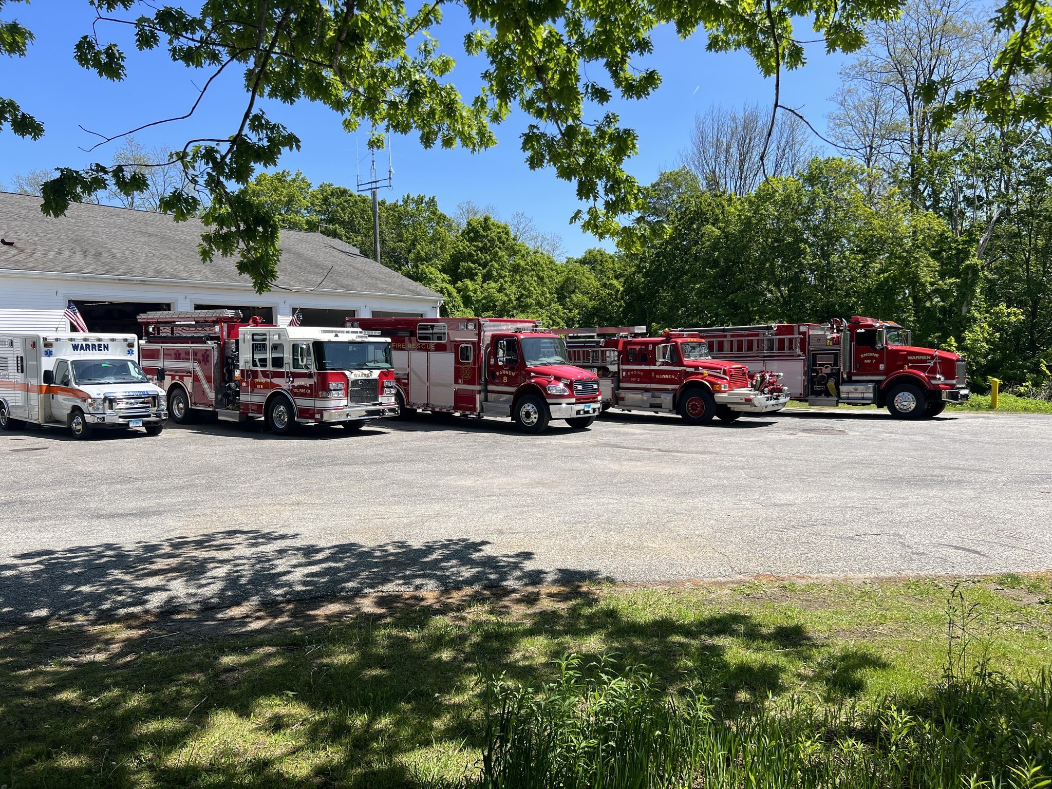 A row of red fire trucks and an ambulance are parked outside a fire station on a sunny day, surrounded by green trees and a clear blue sky.