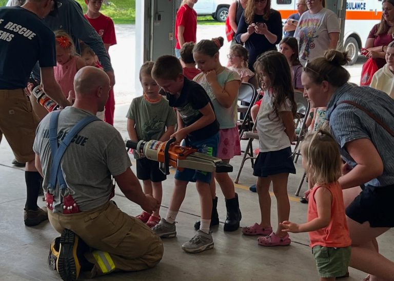 A firefighter kneels on the floor, helping a group of children, one of whom is holding a large rescue tool. Adults and more children watch in a garage with an ambulance in the background.