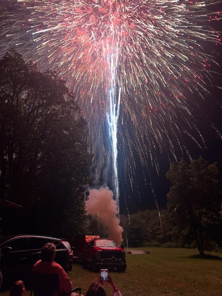 People watch a large, colorful fireworks display lighting up the night sky above trees and parked vehicles, with someone in the foreground taking a photo on their phone. Smoke rises from the fireworks launch point.