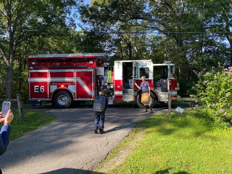 A red fire truck labeled "E6" is parked on a driveway surrounded by trees. A firefighter steps out while a child with a backpack approaches. Another person is taking a photo with a phone.
