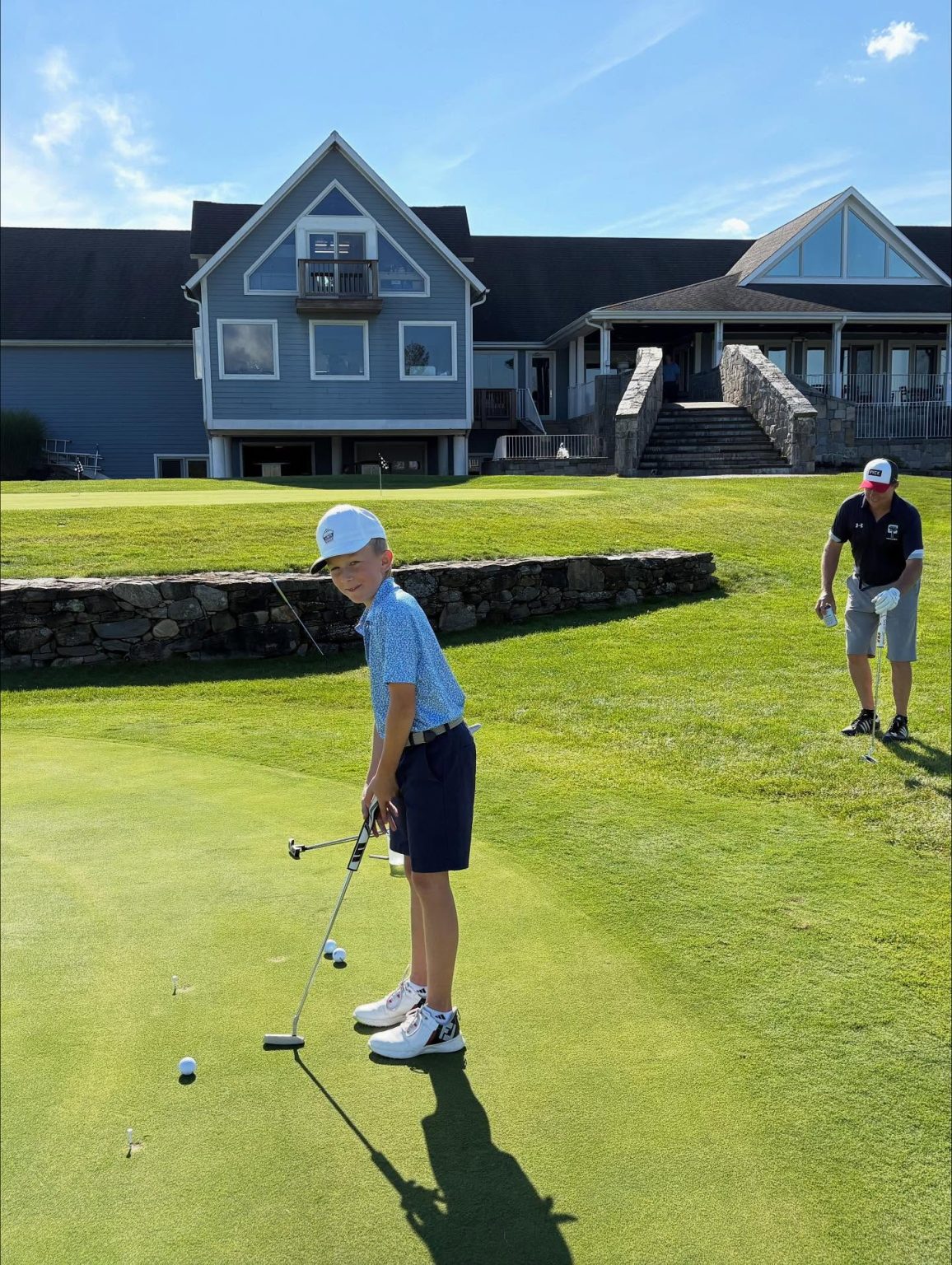 A young boy in golf attire lines up a putt on a green, holding a club. An adult man in a black shirt and shorts stands nearby, preparing for his turn. A stone wall and building are in the background.