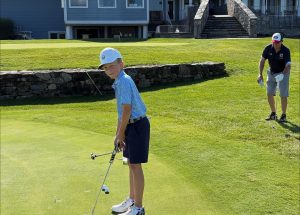 A young boy in golf attire lines up a putt on a green, holding a club. An adult man in a black shirt and shorts stands nearby, preparing for his turn. A stone wall and building are in the background.