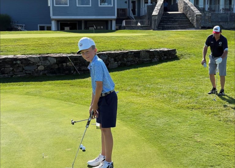 A young boy in golf attire lines up a putt on a green, holding a club. An adult man in a black shirt and shorts stands nearby, preparing for his turn. A stone wall and building are in the background.