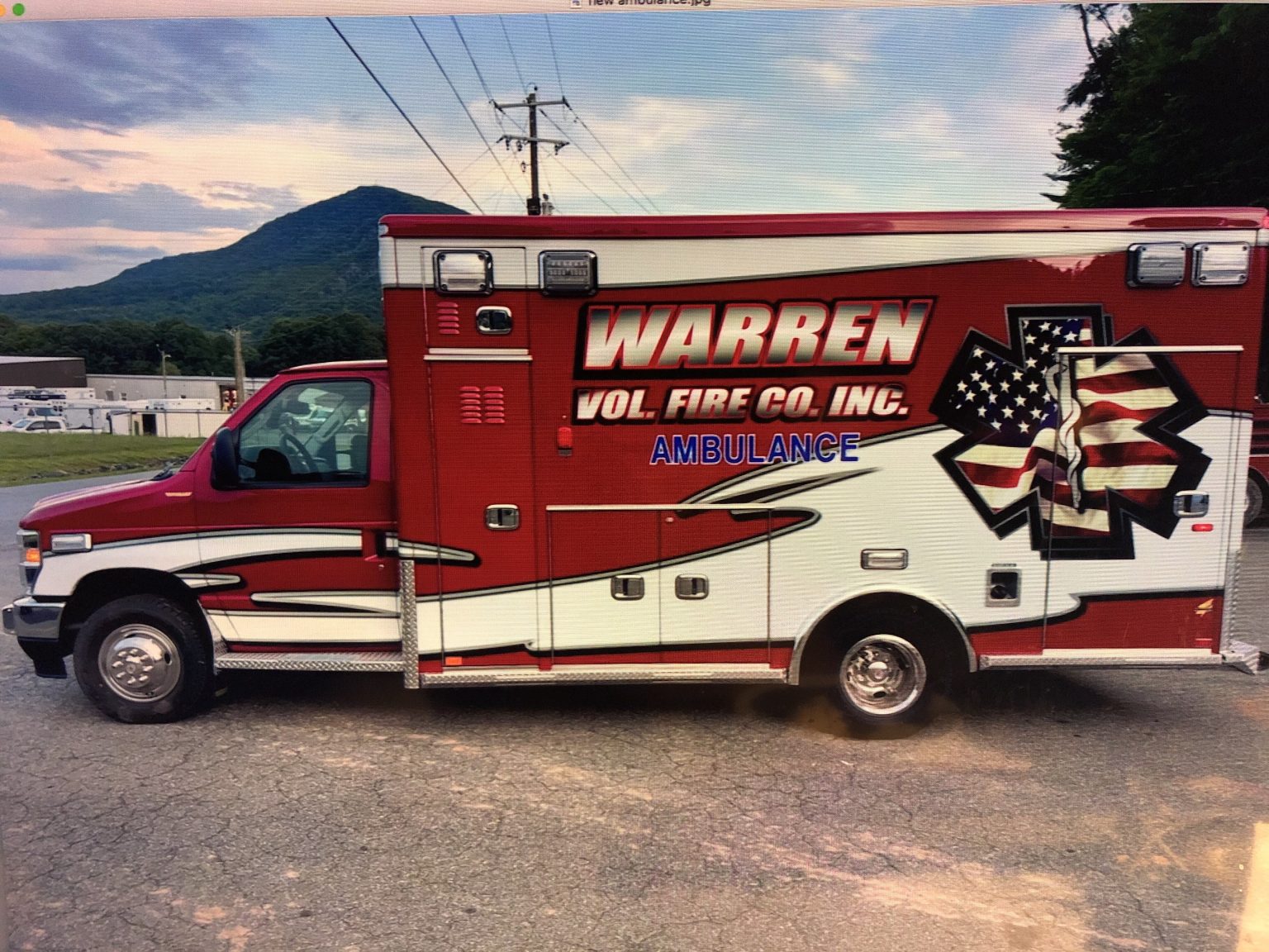 A red and white ambulance marked "Warren Vol. Fire Co. Inc." with an American flag design is parked on a paved surface with trees, a mountain, and buildings in the background under a partly cloudy sky.