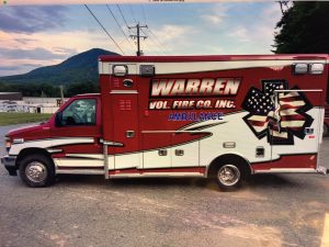 A red and white ambulance marked "Warren Vol. Fire Co. Inc." with an American flag design is parked on a paved surface with trees, a mountain, and buildings in the background under a partly cloudy sky.
