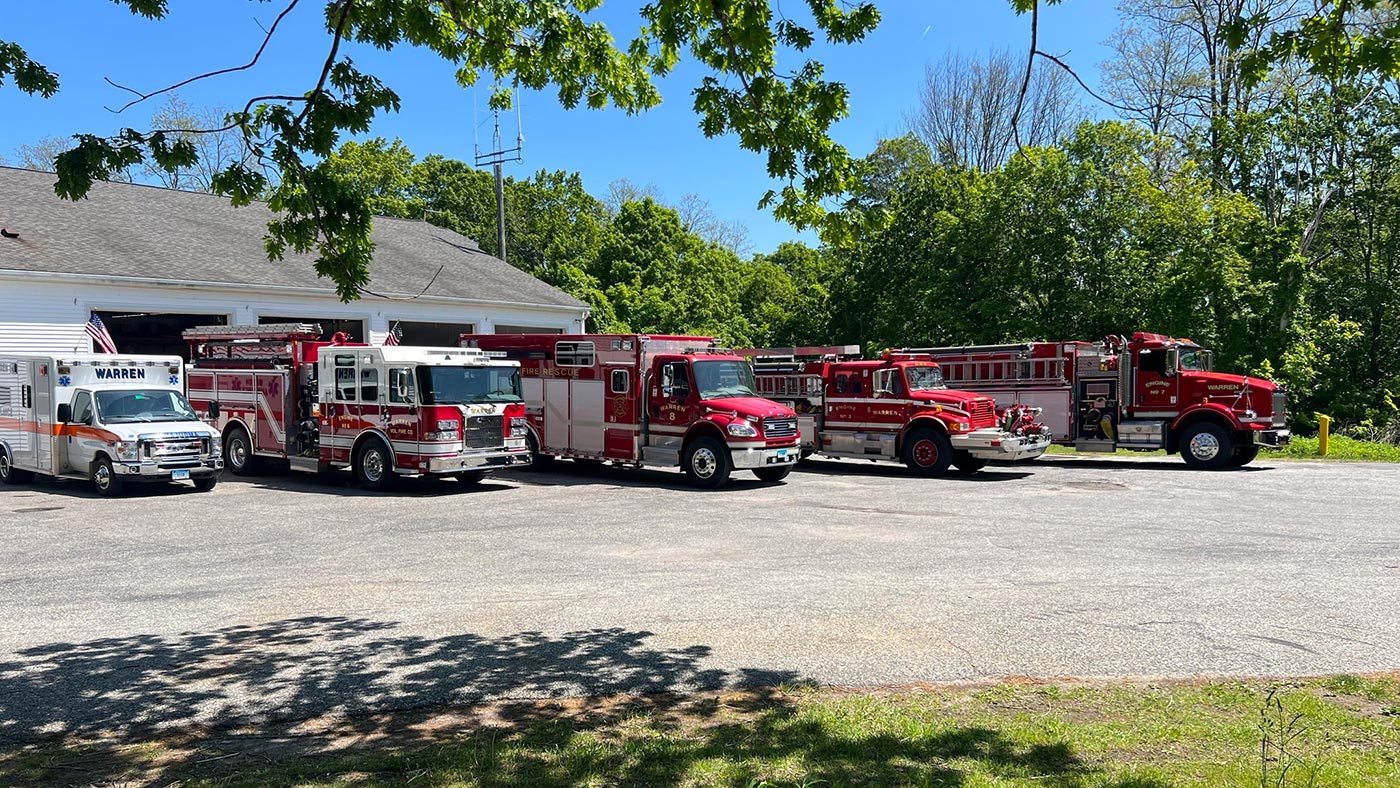 Four red fire trucks and a white emergency vehicle are parked in front of a fire station on a sunny day, surrounded by green trees and a clear blue sky.