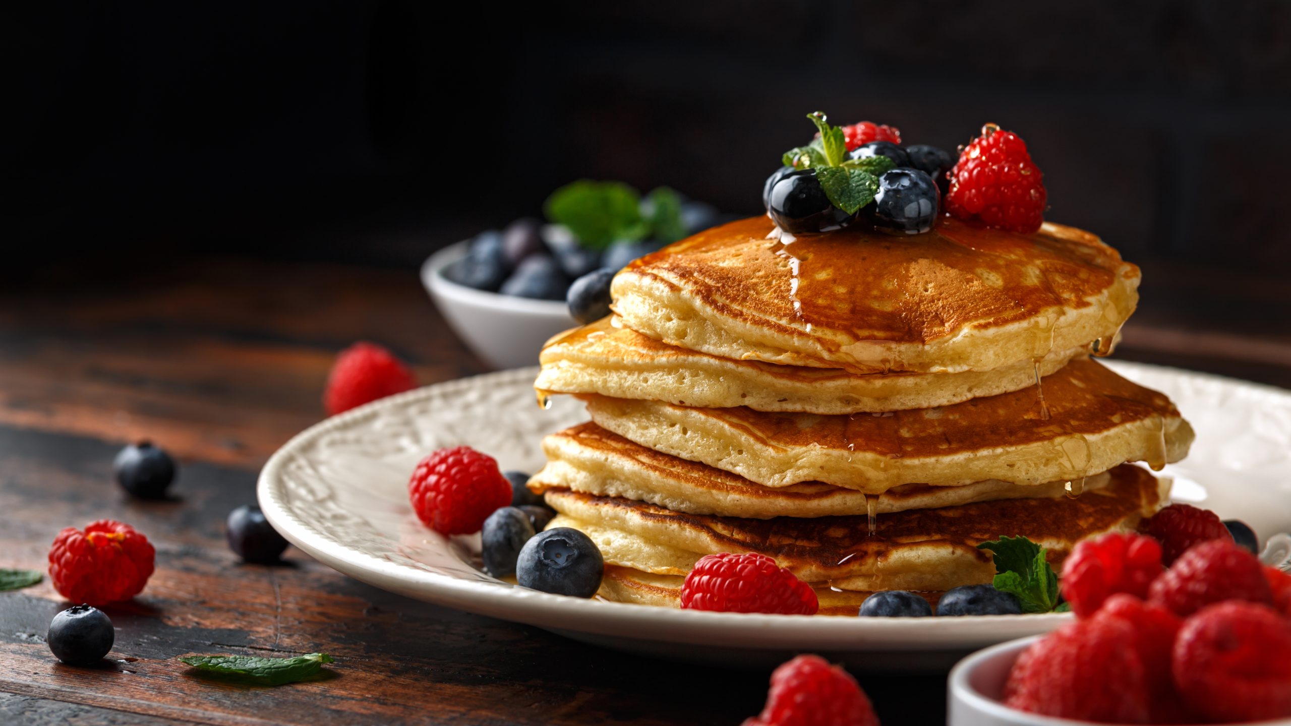 A stack of fluffy pancakes topped with blueberries, raspberries, and mint, drizzled with syrup, sits on a white plate. More fresh berries are scattered around the plate and in small bowls in the background.